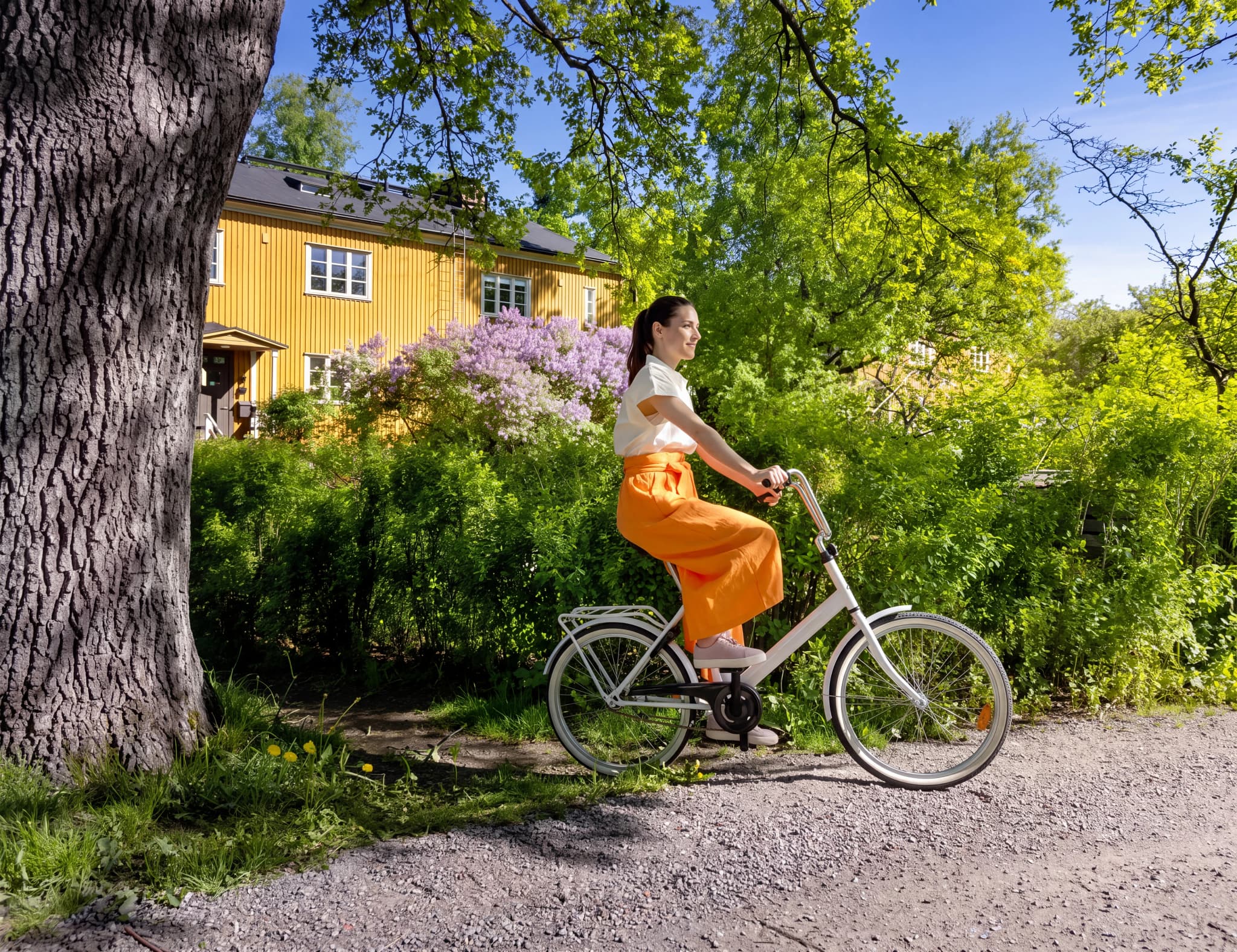 Kvinna cyklar på en grusväg bredvid en gul byggnad och lila blommor.