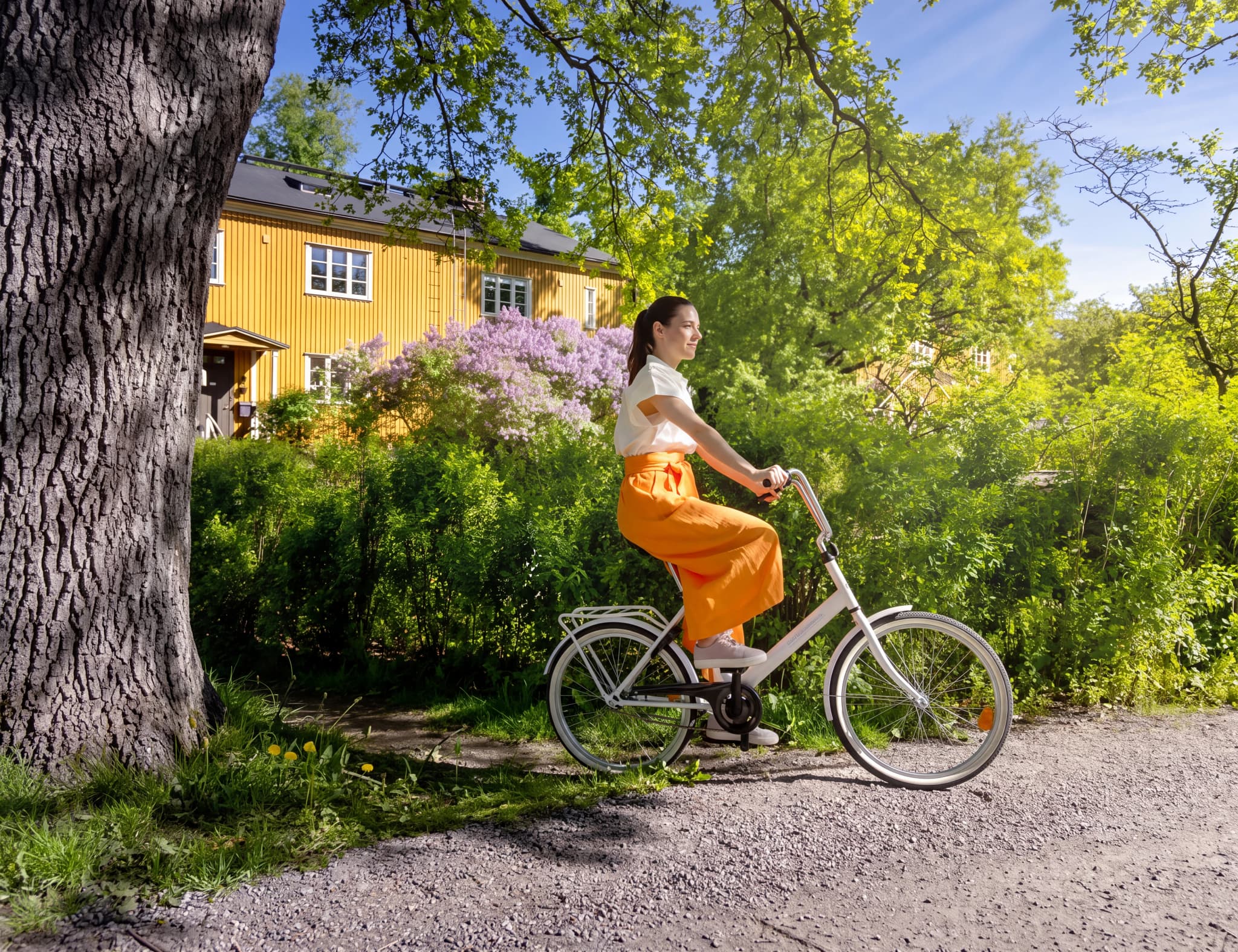 En kvinna i orange kläder cyklar på en grusväg framför ett gult hus med vita fönster och blommande syrener.