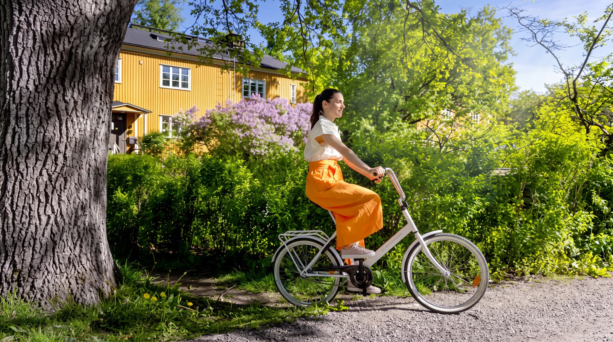 Kvinna cyklar förbi ett gult hus med lila syrenbuskar i en lummig park.