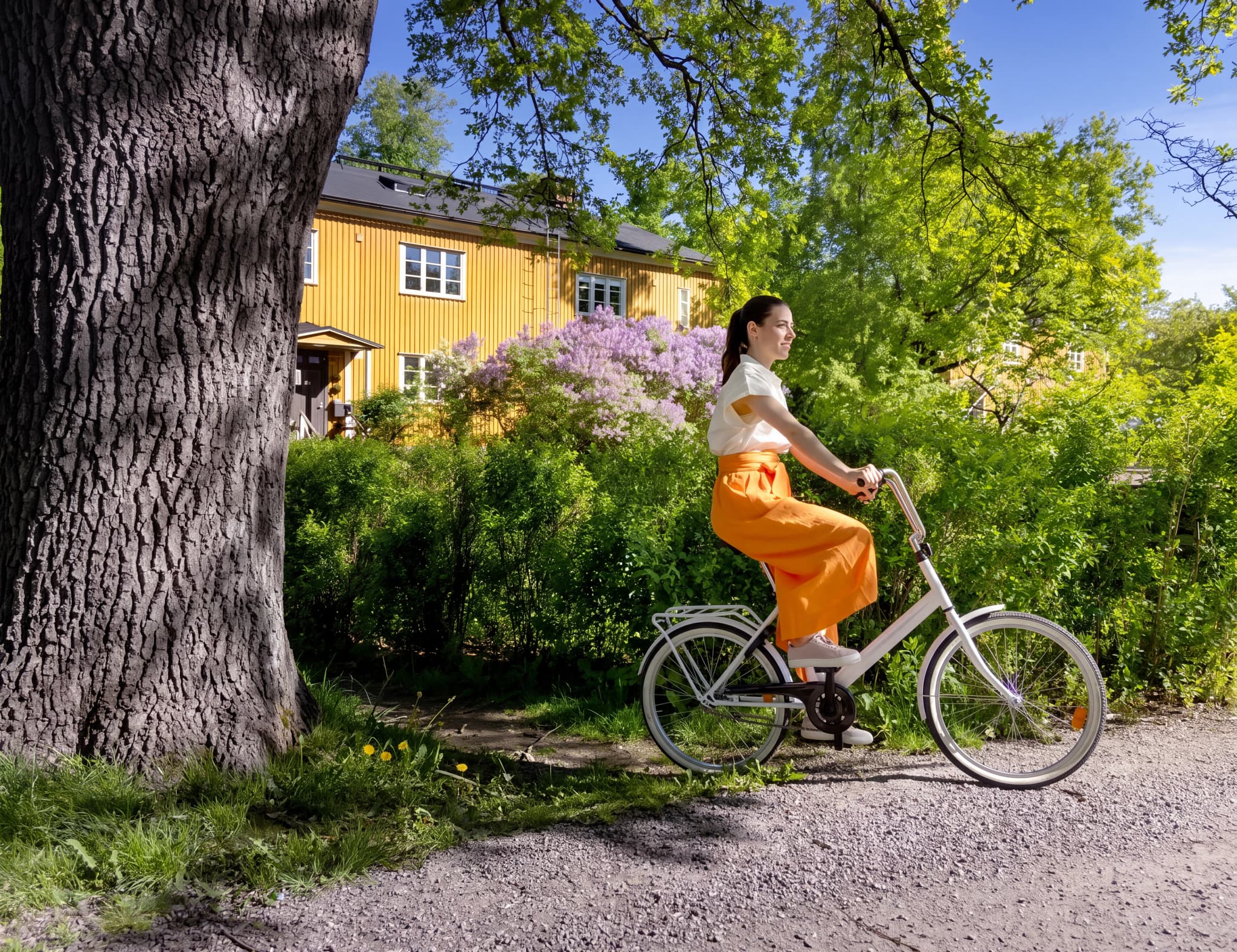Kvinna cyklar på en grusväg genom en lummig park med en gul byggnad i bakgrunden.