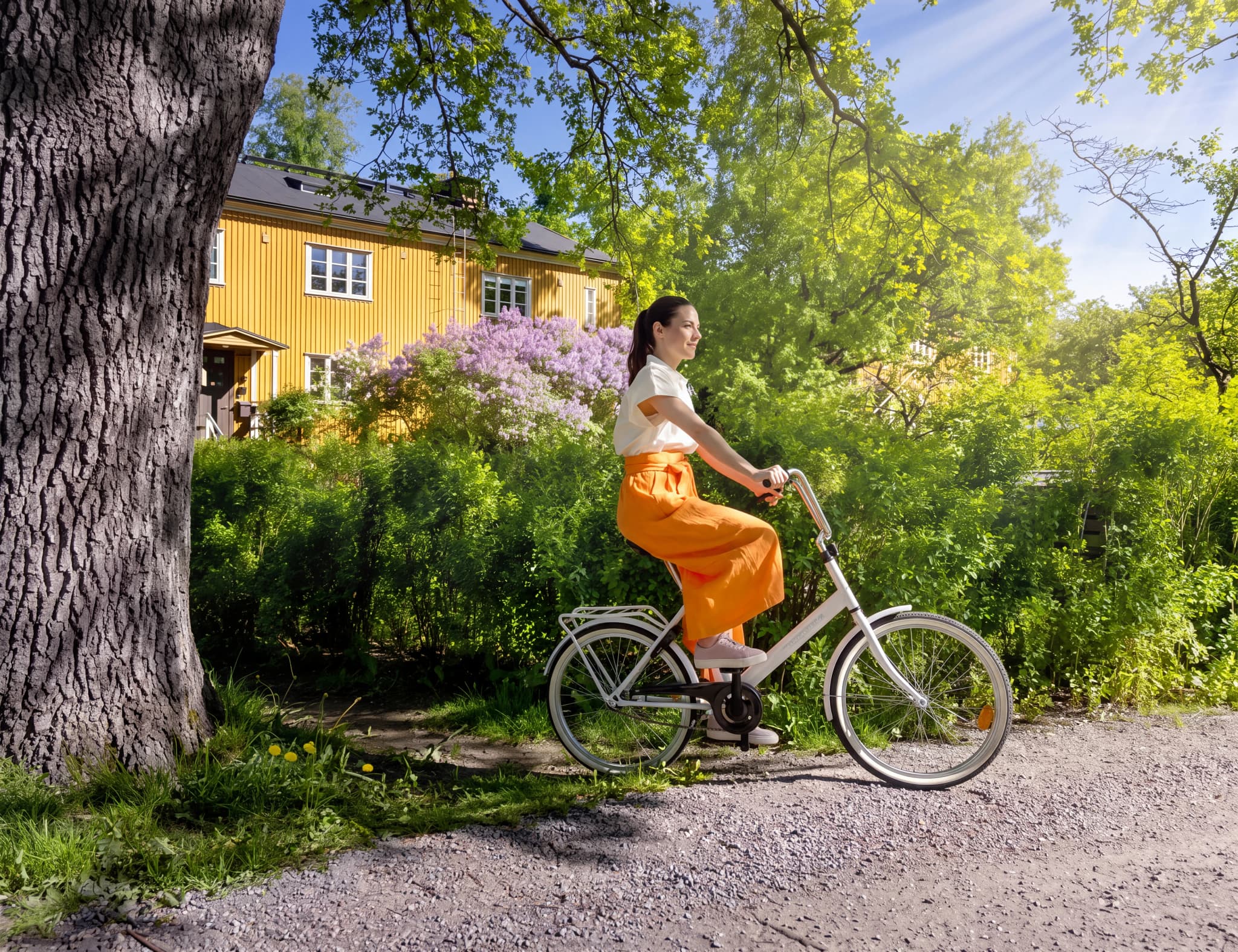 Kvinna cyklar på en grusväg framför ett gult hus med lila syrener.