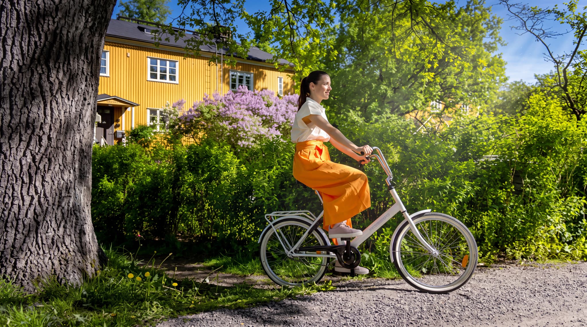 Kvinna cyklar på en grusväg förbi ett gult hus med lila syrener och träd i solsken.