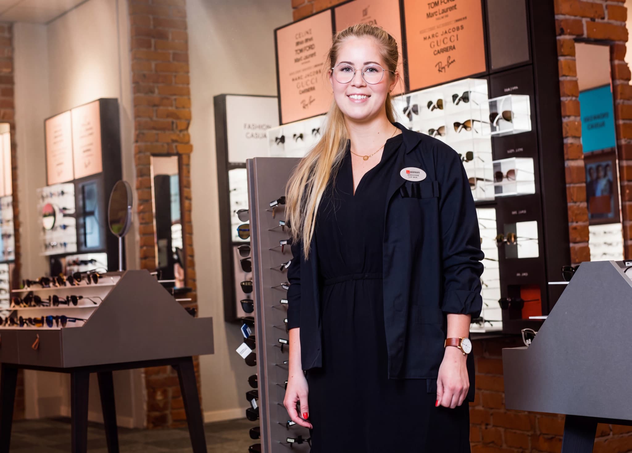 En ung kvinna med glasögon och ett stort leende, klädd i en svart skjorta och klänning, står i en butik som säljer glasögon.