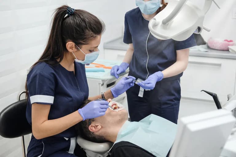 Tandläkare undersöker patientens tänder i en behandlingsstol, med en assistent som håller instrument.