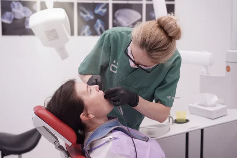 Tandläkare i gröna arbetskläder undersöker en patientens mun med ett instrument i en tandläkarmottagning.