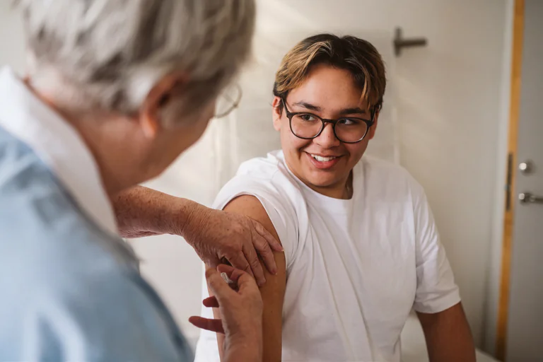 Äldre sjuksköterska ger vaccination till en leende tonåring i glasögon i ett undersökningsrum.