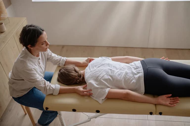 Terapeut masserar klientens nacke och axlar på en massagebänk i ett ljust behandlingsrum.