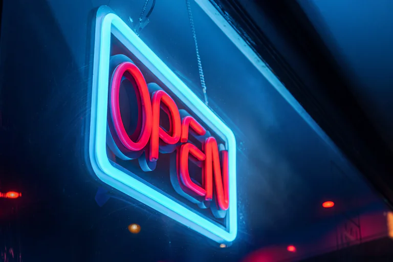 En neonupplyst 'Open'-skylt hänger från ett tak i ett mörkt utrymme.