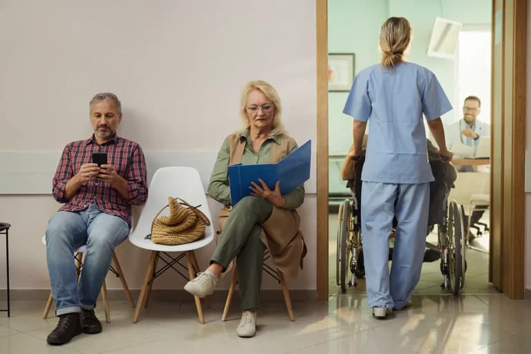 Sjuksköterska skjuter en patient i rullstol genom en korridor i en väntsal, medan två patienter sitter och väntar.