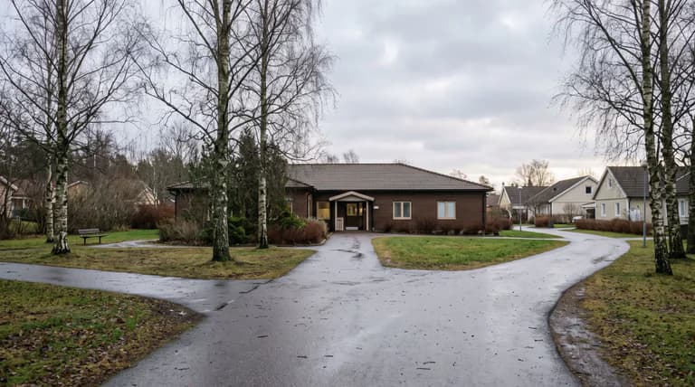 En asfalterad gångväg delar sig i ett vägskäl framför en låg tegelbyggnad omgiven av kala björkar och gräsmattor.