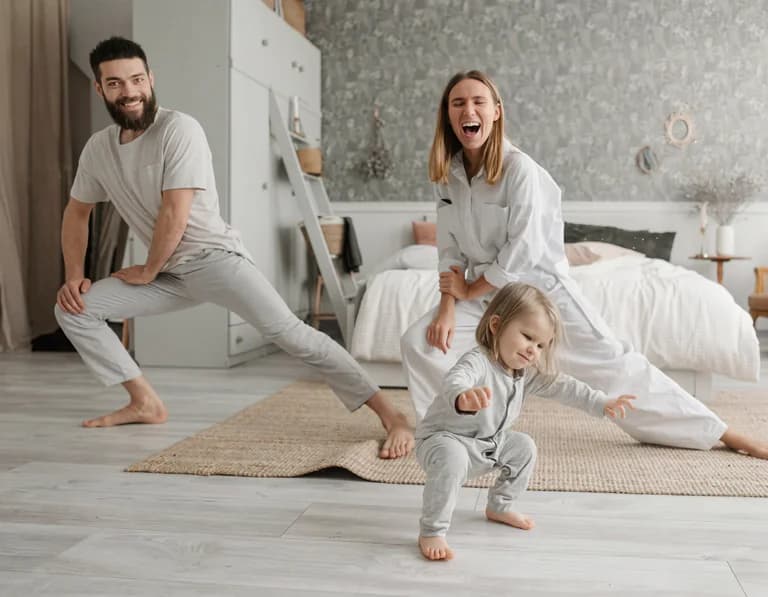 En familj, två vuxna och ett litet barn, stretchar och rör sig på golvet i ett sovrum.