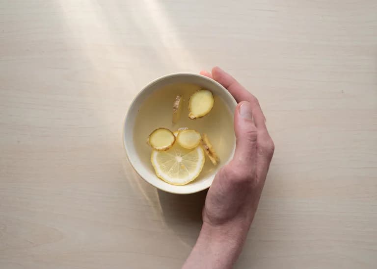 En hand håller en skål med en varm dryck som innehåller ingefära, citronskiva och andra ingredienser.