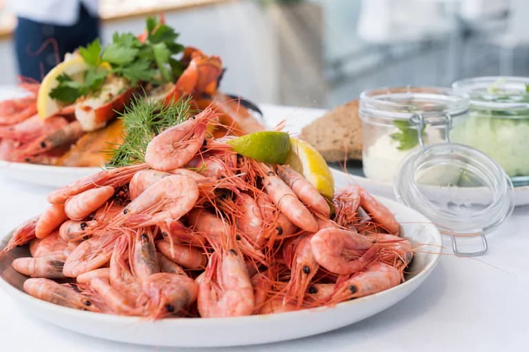 En tallrik med små rosa räkor garnerade med dill och en citronklyfta, med glasburkar med smörgåspålägg och en brödskiva i bakgrunden.