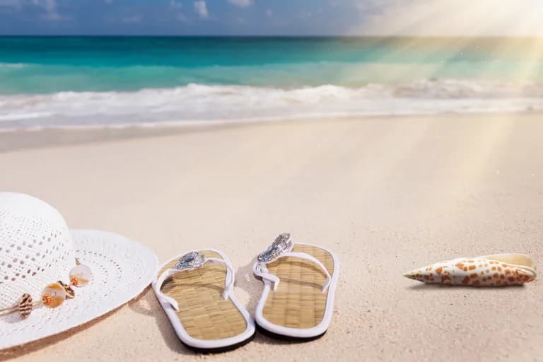 En vit sommarhatt, ett par flip-flops med bling och en snäcka ligger på sanden vid havet.