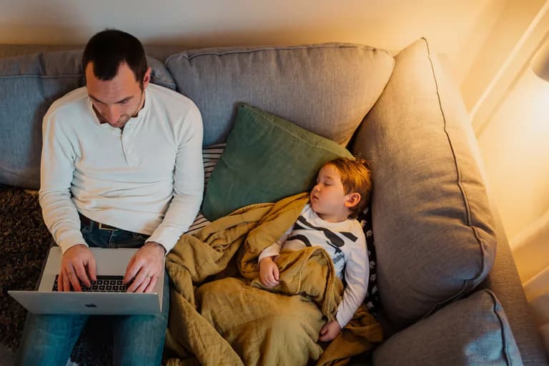 Far arbetar på laptop medan ett barn sover på soffan