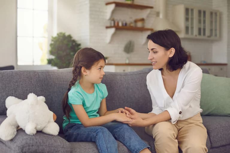 Mor sitter på en soffa och pratar med sin dotter som sitter bredvid henne i ett vardagsrum.