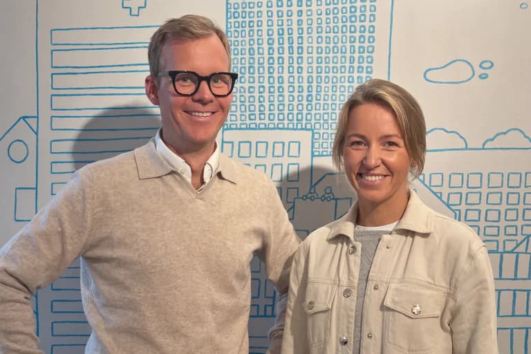 En man och en kvinna ler mot kameran framför en väggmålning som föreställer en stadssiluett med byggnader.