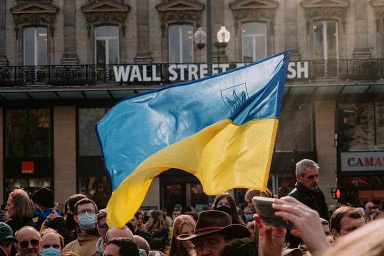 En grupp människor samlas utomhus med en stor ukrainsk flagga som viftas framför en byggnad med texten "WALL STREET". Några personer bär munskydd.