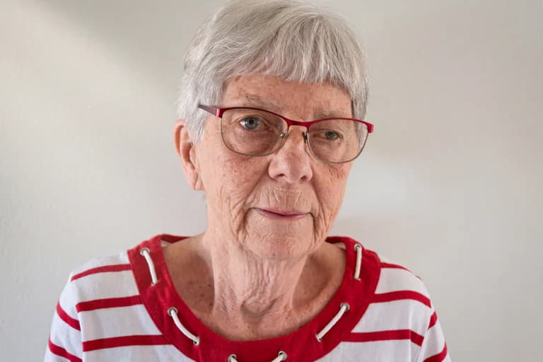 Äldre kvinna med grått hår och glasögon bär en randig tröja med röd kant och snörning.