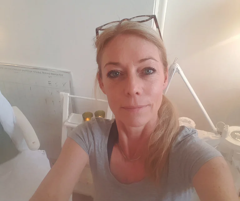 Kvinna med glasögon på huvudet i ett rum med vit vägg och hylla.