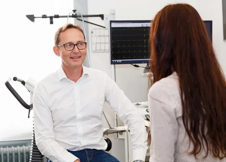 Läkare i vit skjorta ler och pratar med en patient vars hår syns på höger sida, med en hjärtmonitor i bakgrunden.