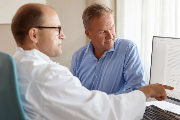 Läkare i vit rock pekar på datorskärmen och förklarar resultat för patient i blå skjorta.