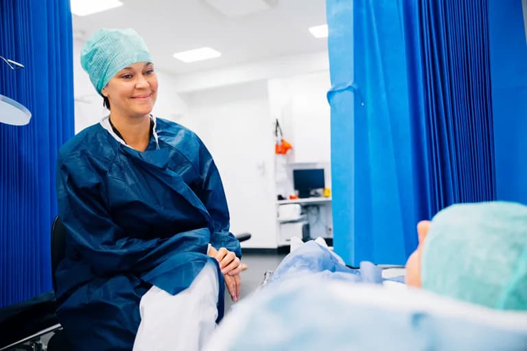 En sjuksköterska i operationskläder sitter och pratar med en patient som ligger i en sjukhussäng, omgiven av blåa draperier i en klinisk miljö.