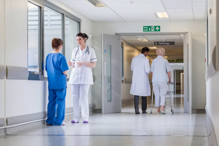 Två sjuksköterskor och en läkare konverserar i en sjukhuskorridor med en patient som går förbi med en rollator.