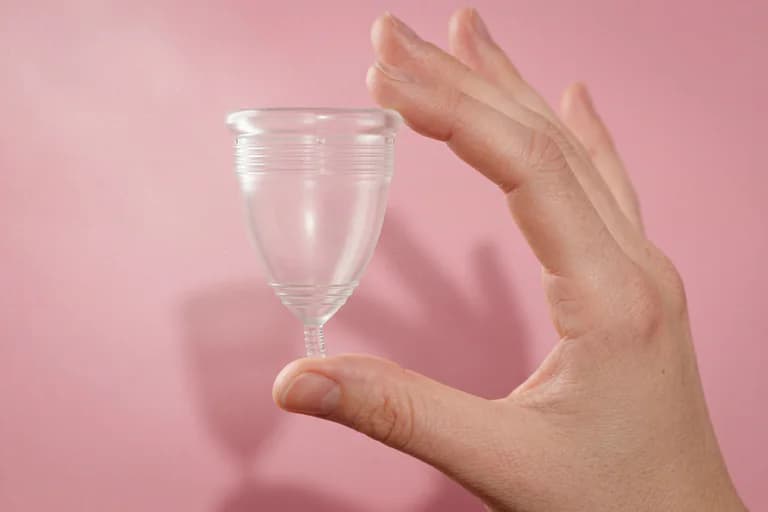 En hand som håller en vit menstruationskopp mot en rosa bakgrund.