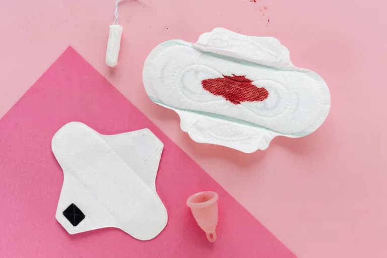 En platt överbild av olika mensskydd, inklusive en tampong, ett binda med en blodfläck, en tygbinda och en menskopp, arrangerade på en rosa bakgrund.